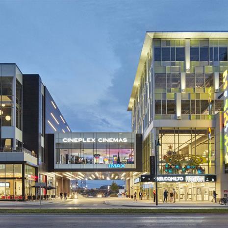 two midrise retail and office mixd-use buildings lit up at dusk, connected with an elevated pedestrian walkway over a road