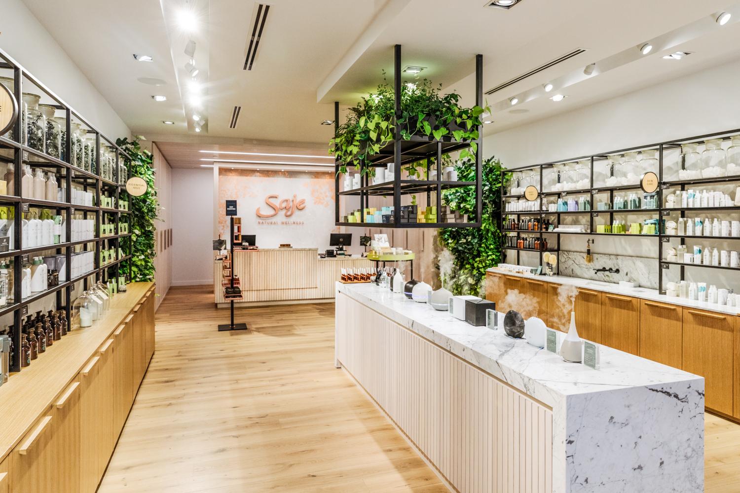 inside a Saje store with a white marble display coutner of diffusers in the middle, a hanging planter and two living walls with greenery and products lining both walls