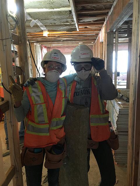 two women wearing hard hats and safety vests with their arms around each other's shoulders on a construction site