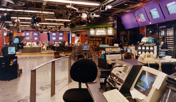 broadcast newsroom in the year 2000 with computers, tv screens and professional lighting, and a view of an urban streetfront in the background