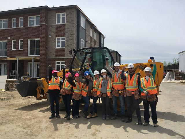 9 people in hard hats and safety vests raise their fists while standing in front of a backhoe