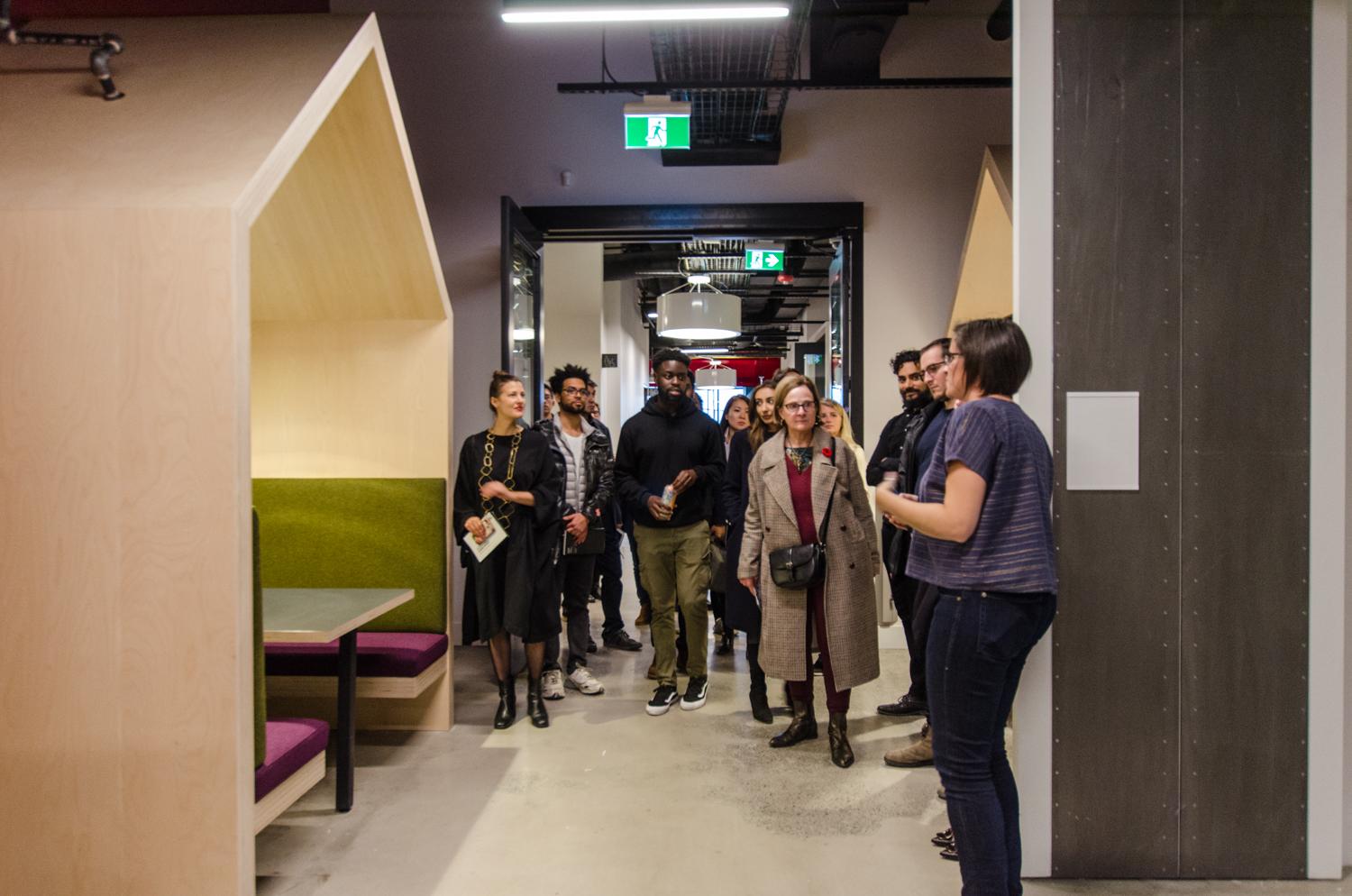 Tor McGlade leading an interior design tour at Artscape Daniels Launchpad, showing the group a covered wooden seating nook with olive and plum cushions