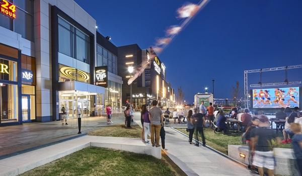 people in a public realm with an outdoor screening underway next to a Cineplex and retail and entertainment block