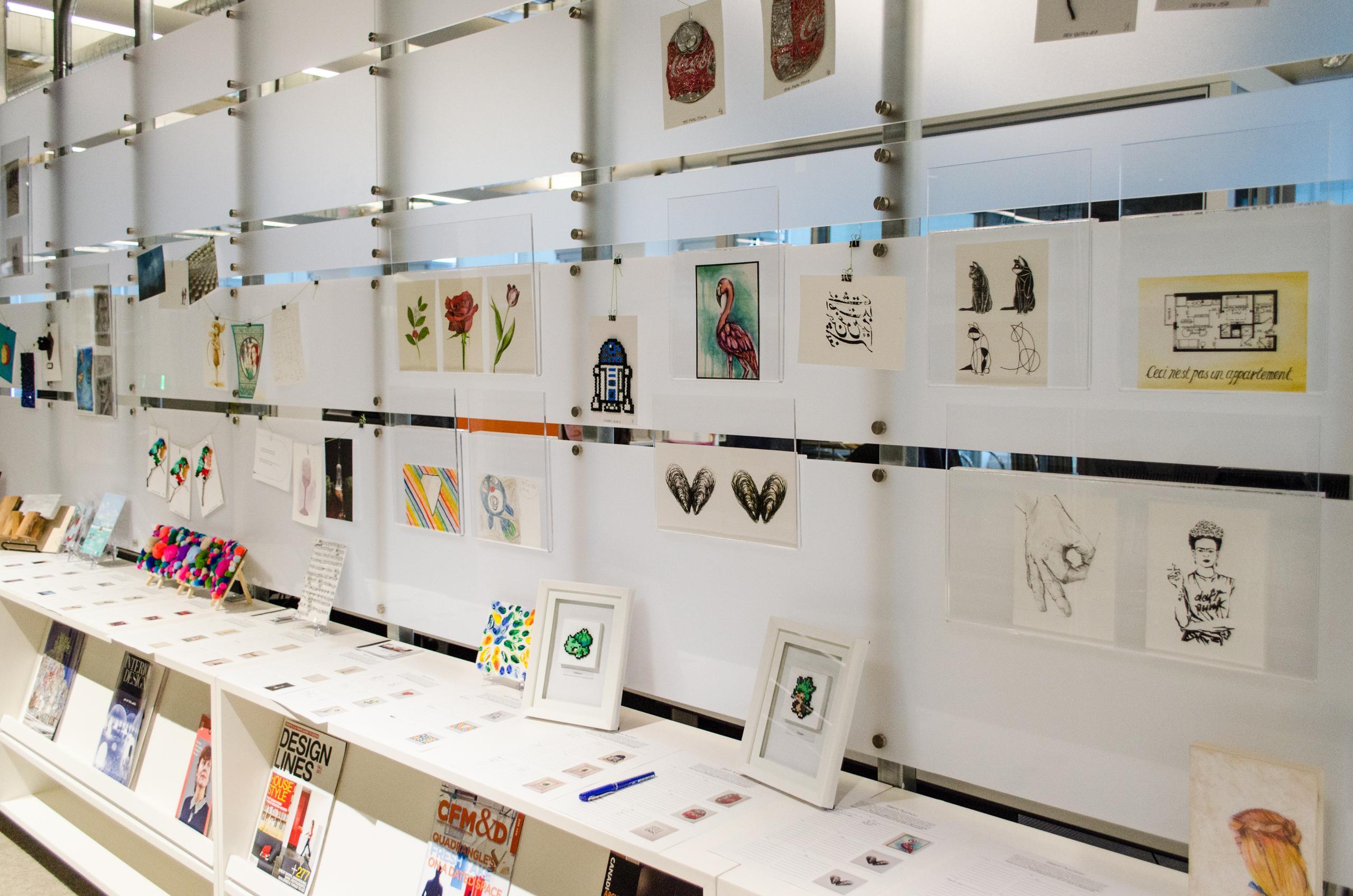 lots of postcard sized artwork hung up on a wall display in the Quadrangle studio alongside silent auction bid sheets