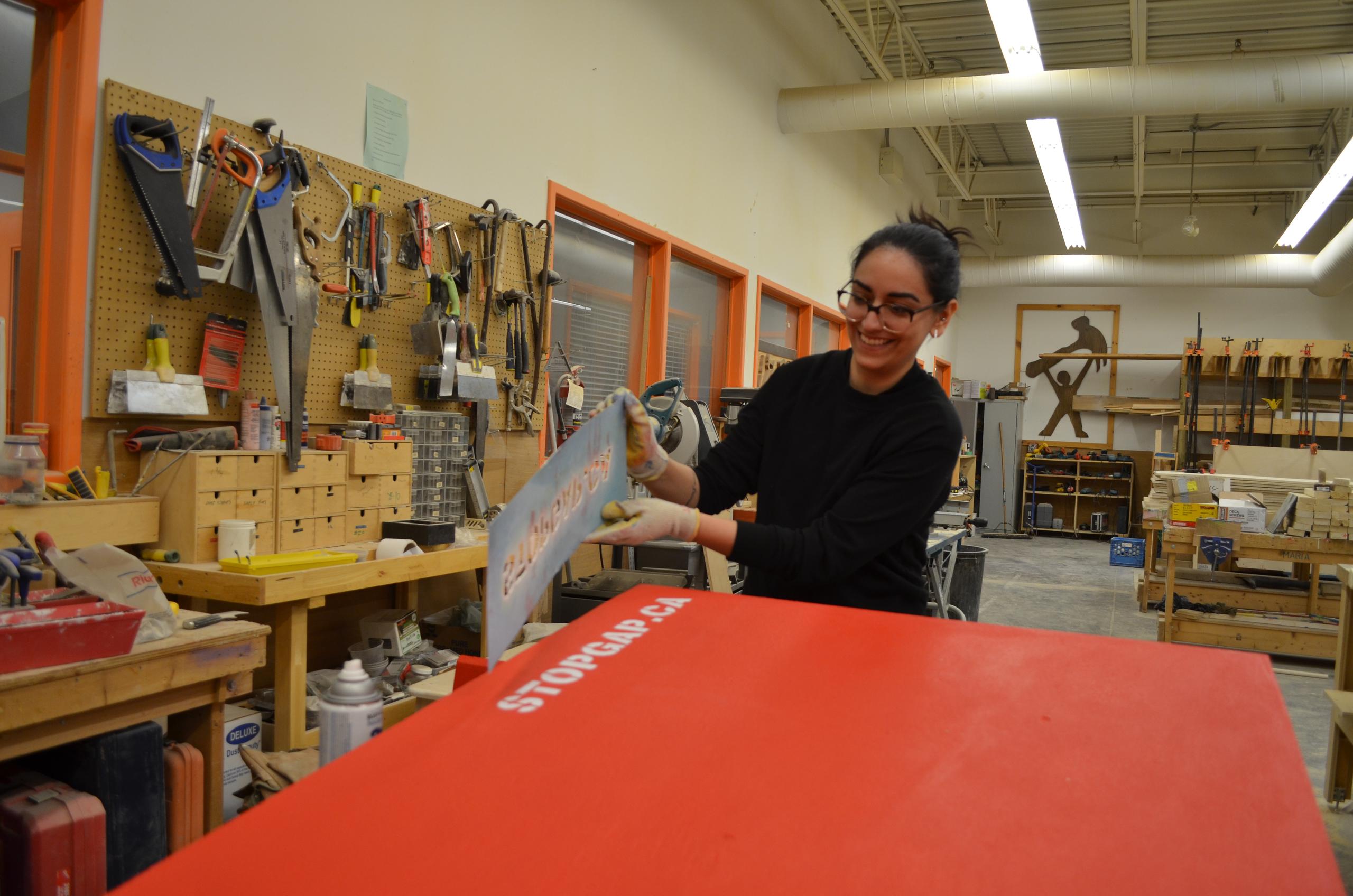 a woman smiles as she removes the STOPGAP.CA stencil from a freshly painted red ramp