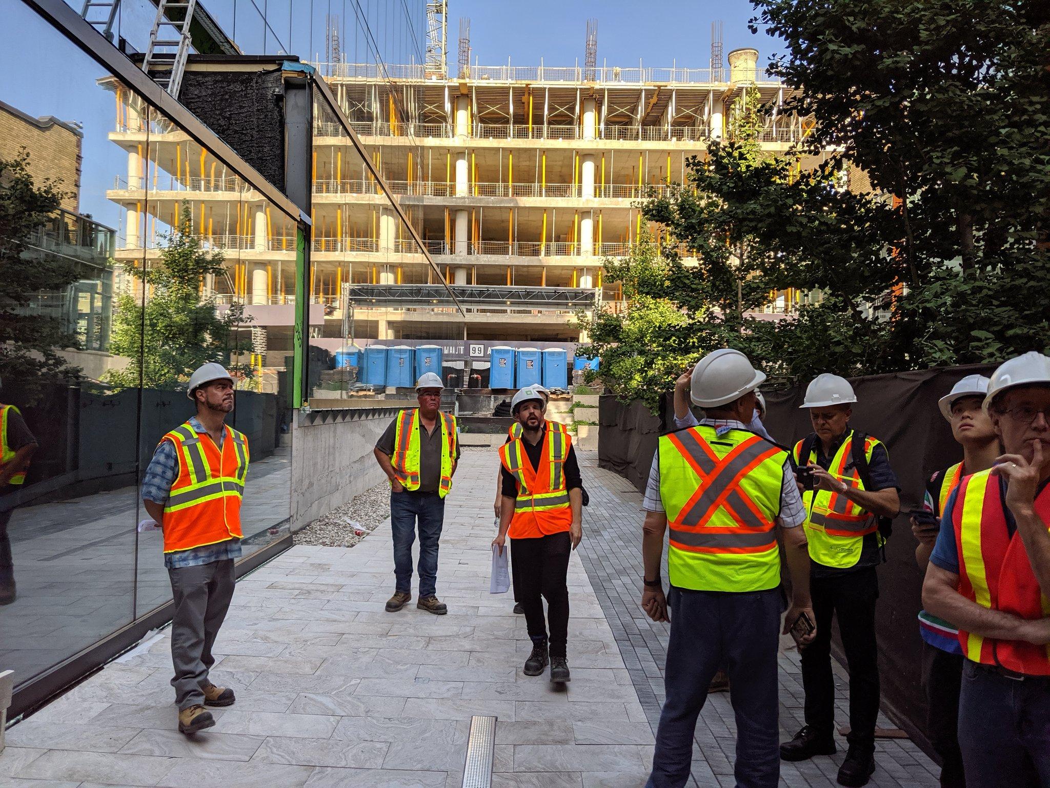 Wayne McMillan walking down a laneway with other people in construction site safety gear, with the half-finished 80 Alantic in the background showing several floors of exposed wood ceiling and columns