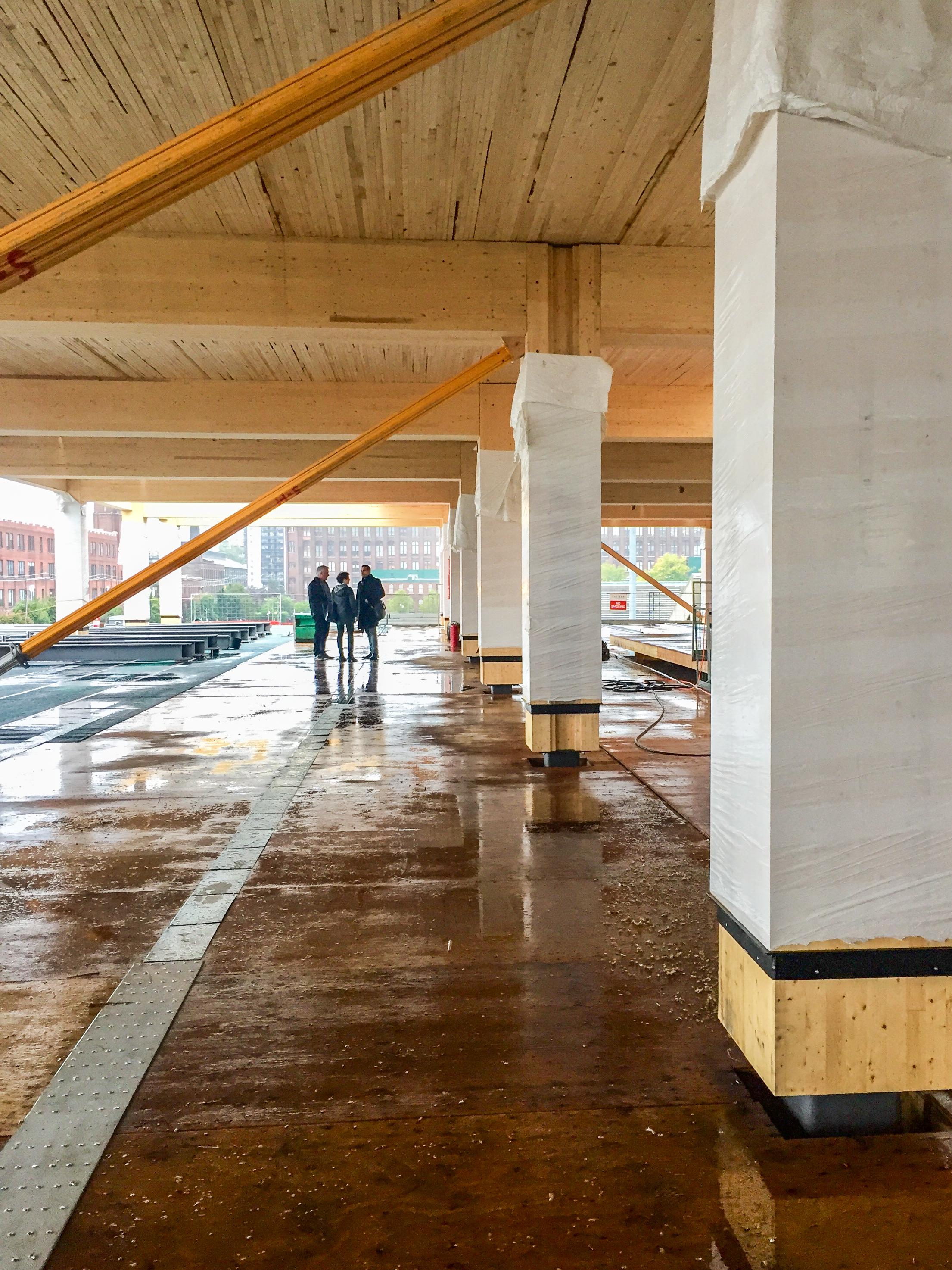 three people in the distance standing in the under construction 80 Atlantic looking at the wood post and beams and NLT deck timber ceiling, temporary braces are installed at the post and beams