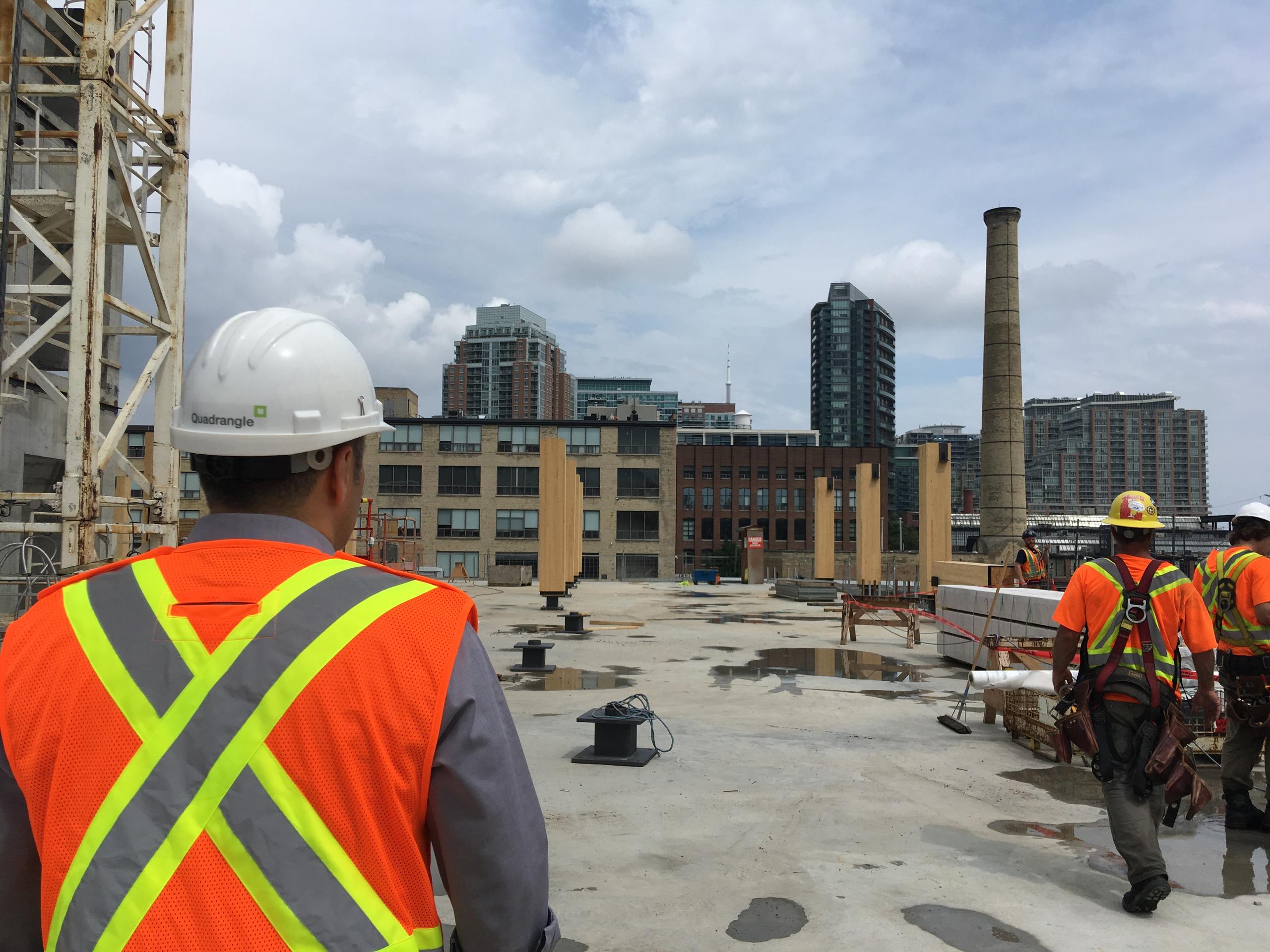 Richard Witt seen from behind wearing safety vest and Quadrangle branded hard hat in the foreground on site at 80 Atlantic looking at upright glulam columns and Liberty Village condos and heritage smokestack in the distance