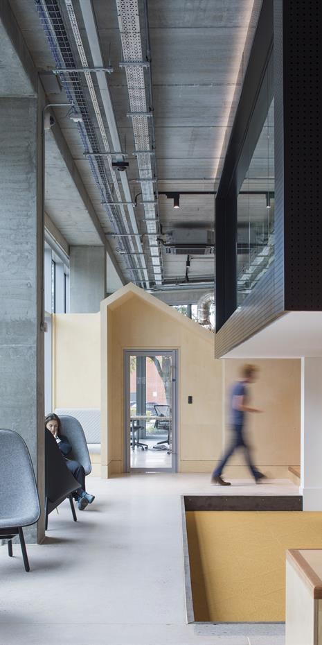 office interior showing seating along a window, wooden wall with doorway, cantilevered black block meeting room with seating underneath