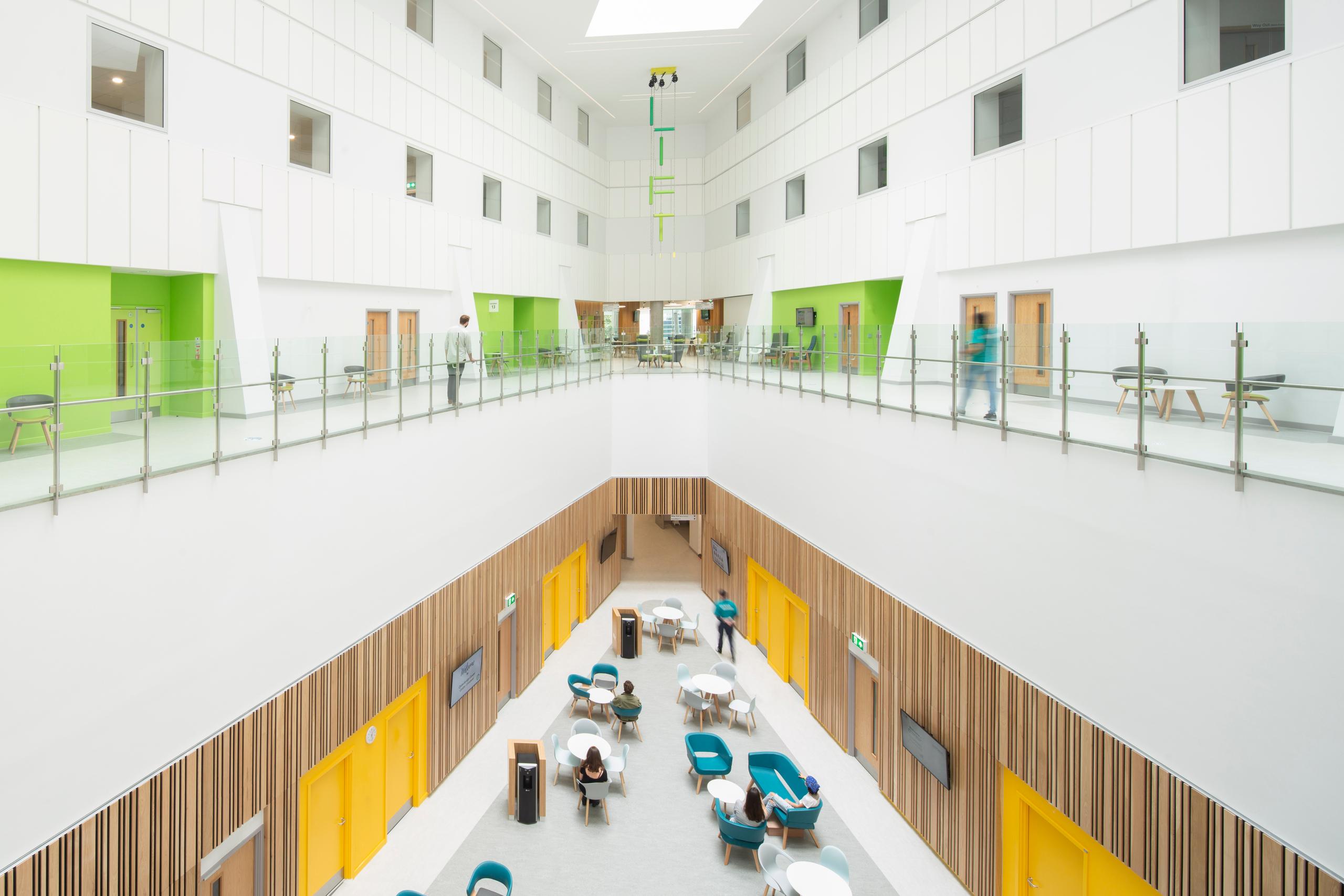 overlooking an atrium inside the Clatterbridge Cancer Centre which has wooden walls and yellow doorways on the lower level and crisp white walls and lime green doorways on the second level with skylights overhead