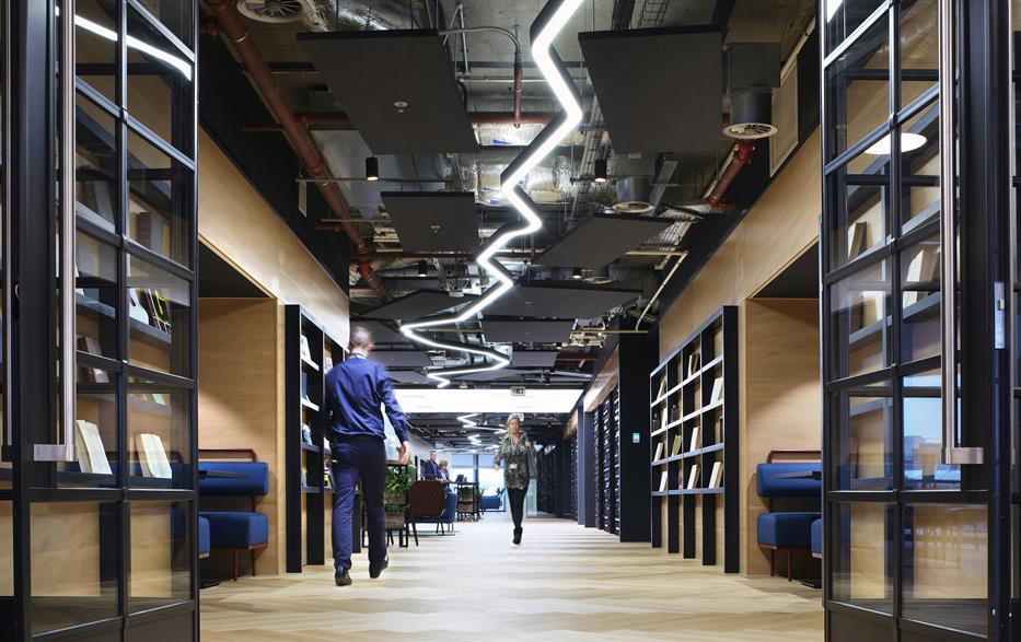 hallway in a modern office with blue bench seating, built in black shelving lining the light coloured wood walls, and a zig zag custom light fixture that runs the length of the hallway